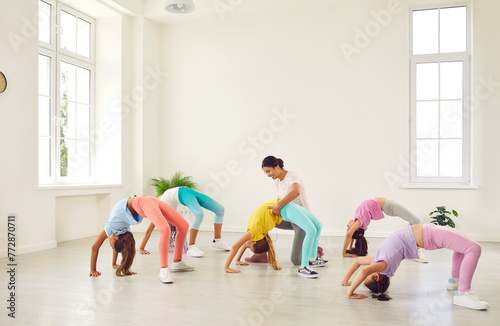 Kids doing gymnastics. Group of children doing gymnastic exercises at a sports school. Female trainer helps little flexible sporty gymnast girls who are doing a bridge position all together photo