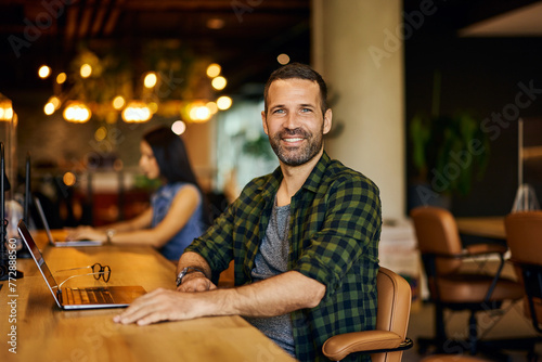 Portrait of a smiling businessman working online in the co-working space, his female colleague working in the background.