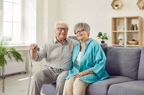 Portrait of a happy senior family couple wife and husband with crutch sitting on sofa in the living room at home. Two smiling gray-haired retired man and woman enjoying time together on retirement.