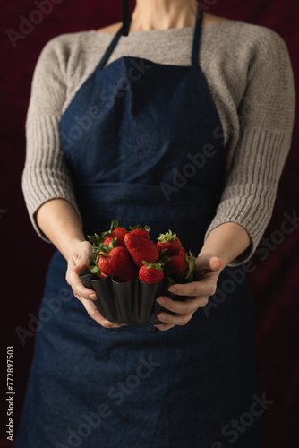 A woman holding a metallic bowl filled with ripe, red strawberries ready to eat, set against a black background