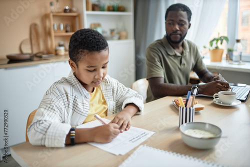 Portrait of young African American boy doing homework at kitchen table with father watching