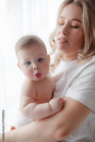A 5-month-old baby and his beautiful blonde mom relax, play and laugh in bed in the bedroom. People a dressed in light home clothes, the family chooses natural textiles