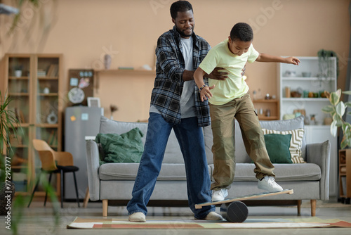 Full length portrait of happy African American father teaching boy skateboarding standing on balance board