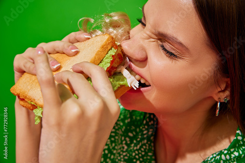 Young woman eating quirky sandwich with doll against red background. Weirdness and surrealism. Close-up. Concept of food pop art photography, creativity, quirky style