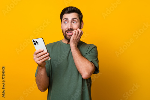 Photo portrait of handsome young guy hold gadget bite nails frightened dressed stylish khaki outfit isolated on yellow color background photo