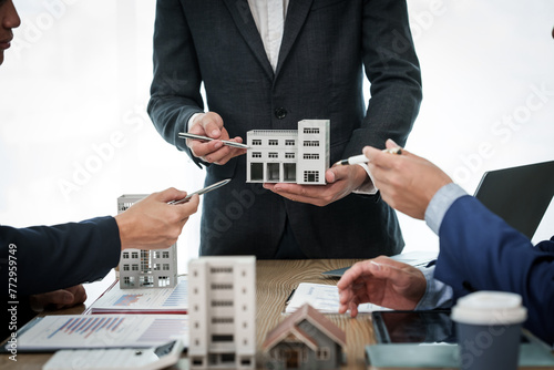 A professional businessman in a suit conducts a meeting at a desk while showcasing a model of a condominium or apartment building, discussing real estate development plans.