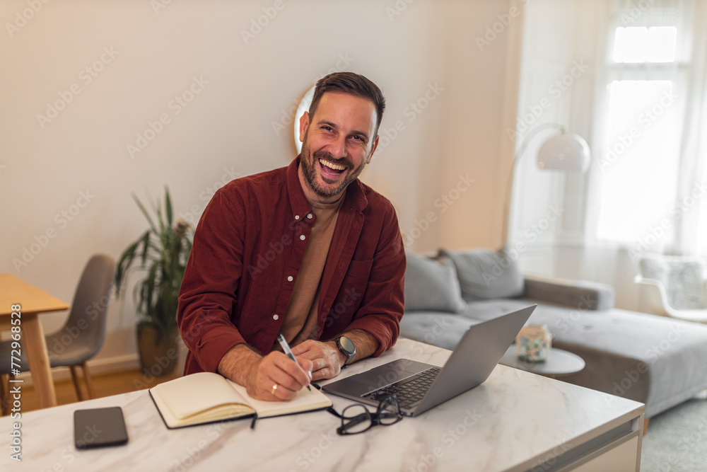 A portrait of a cheerful male entrepreneur working from home and taking notes in front of a laptop A portrait of a cheerful male entrepreneur working from home and taking notes in front of a laptop