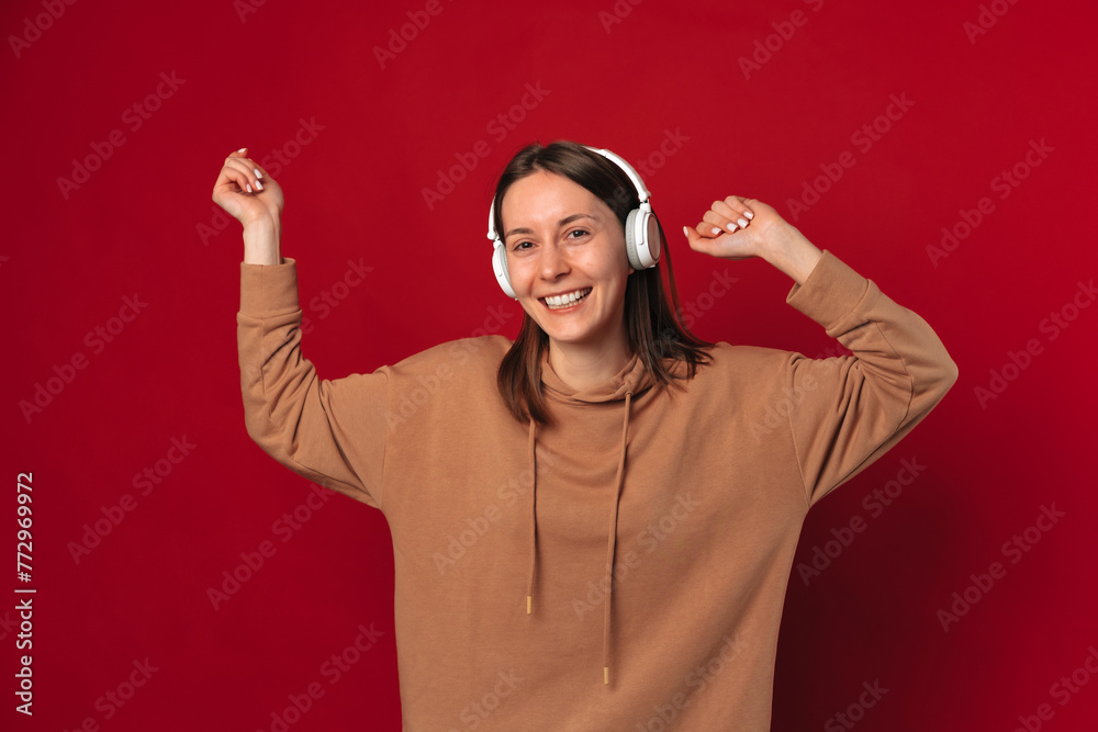 Cheerful young woman wearing headphones is dancing in a studio over red background. Cheerful young woman wearing headphones is dancing in a studio over red background.