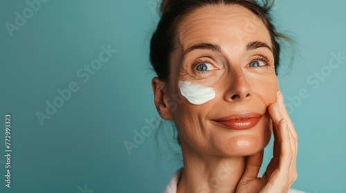 A 50-year-old woman smilingly applies cream to her face with her hand photo