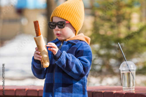 A little boy eats fast food outside in the spring. He will rub the baby in a fashionable plaid shirt, yellow hat and sunglasses. The child eats a hot dog and drinks lemonade.