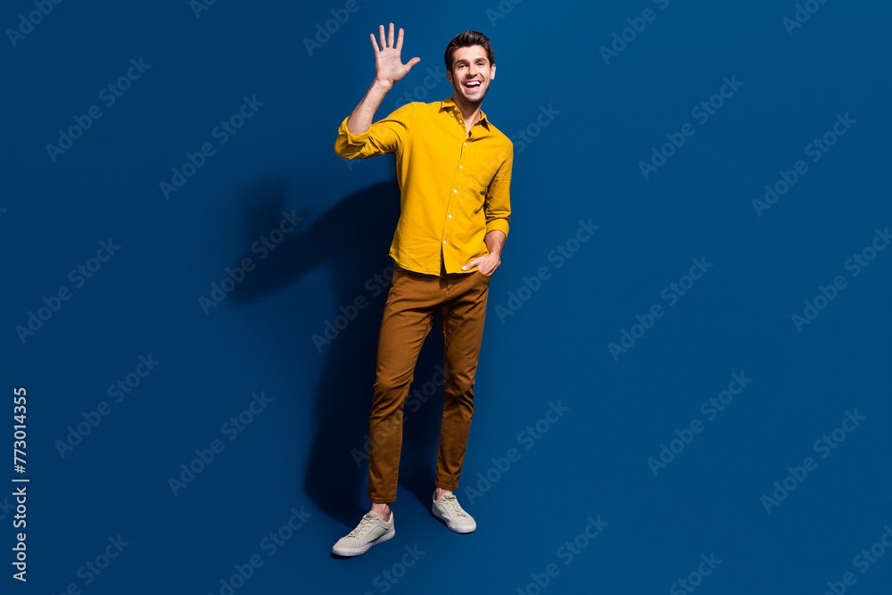Full length photo of nice young male wave hand say excited hello dressed stylish yellow garment isolated on dark blue color background