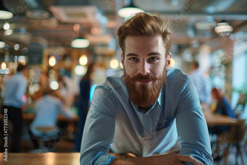 engaging photo of the confidence of a young bearded businessman posing on a desk in a startup office, with a blurred background of energetic employees collaborating and brainstormi