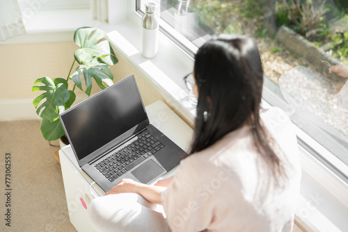 Shot from behind of a woman wearing glasses and a pink top sitting cross-legged on a white chest while watching content on her grey laptop computer and white earphones next to a big bay window