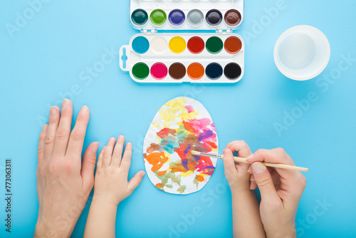 Mother and baby boy hands holding paintbrush and painting colorful egg on paper with watercolor on blue table background. Closeup. Child making easter decoration. Point of view shot. Top down view.