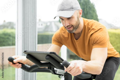 Young Hispanic man with a stubble wearing a grey cap and orange top exercising on a spinning bike in a conservatory while following an online fitness workout on his tablet resting on the handlebar