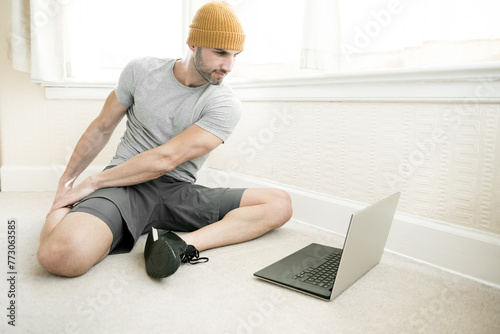 Hispanic young man wearing a yellow hat and grey top stretching his legs on a carpeted floor of a house in Edinburgh, Scotland, UK, as he follows an online workout class on his laptop