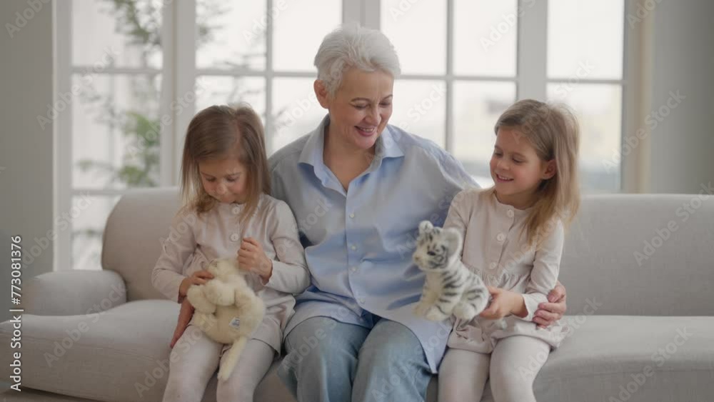Grandmother with granddaughters resting. Happy relaxed older adult granny embracing playing with toys with children twins grandchildren leisure rest on couch at home. Family communication concept. Grandmother with granddaughters resting. Happy relaxed older adult granny embracing playing with toys with children twins grandchildren leisure rest on couch at home. Family communication concept.