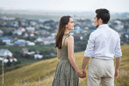 Happy couple of young man and woman smiles holding hands on hill top in cloudy weather. Lovers joy and landscape against distant village