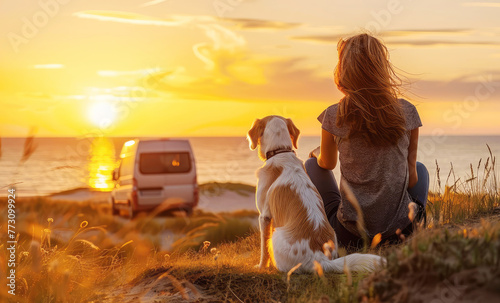 young woman with a dog sits on the coast next to a campervan and enjoys the sunset and evening breeze