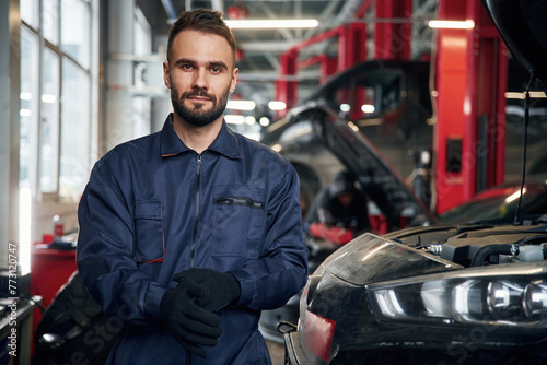 Wearing the gloves, standing. Car repairman is in the garage with automobile