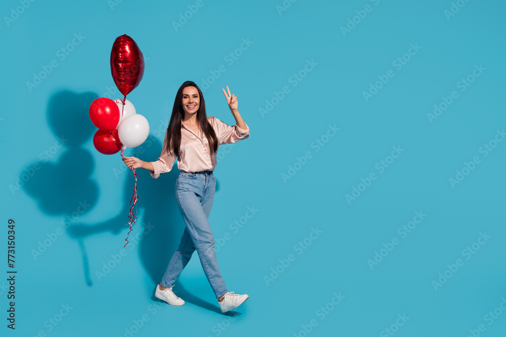 Fototapeta premium Full length photo of lovely girl wear silk blouse hold heart balloons go empty space show v-sign isolated on blue color background