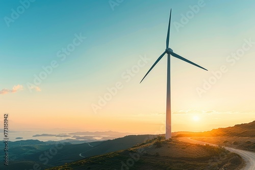 A wind turbine stands atop a hill at sunset, with the sun setting in the background against a clear blue sky