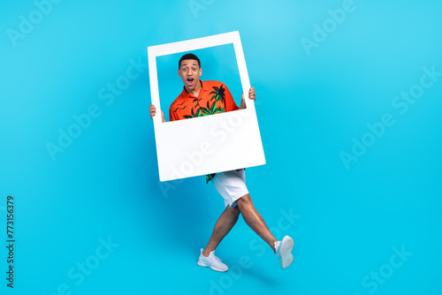 Full length photo of good mood excited guy dressed print shirt holding paper photo walking isolated blue color background