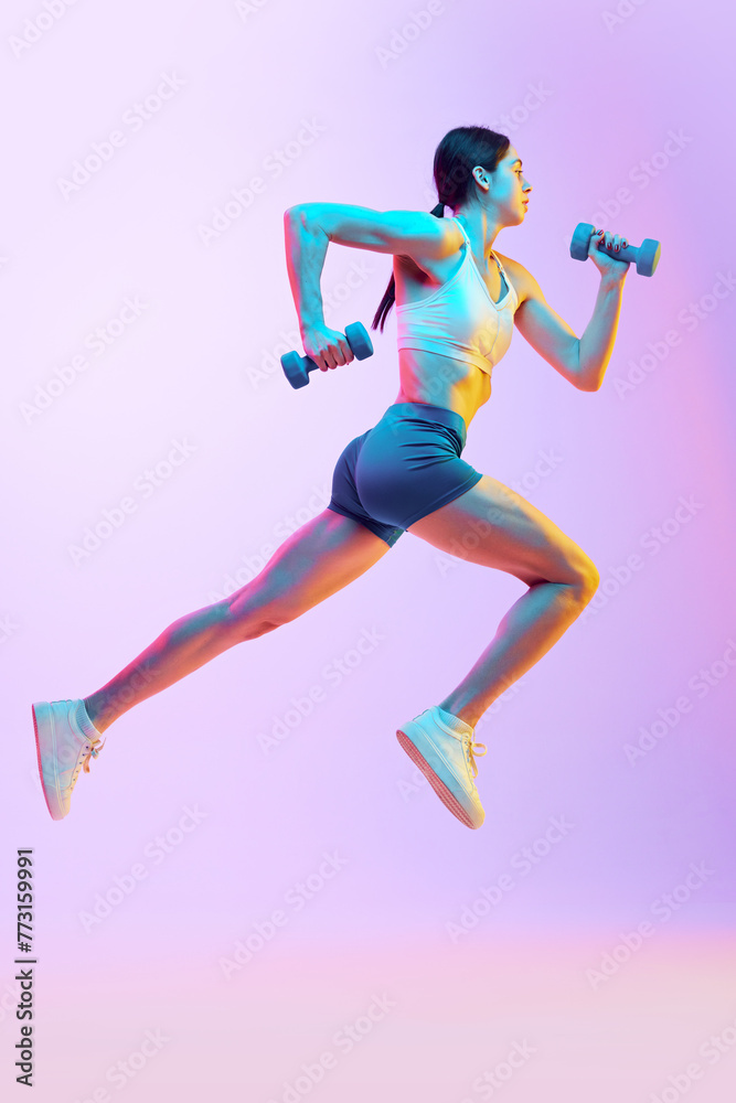 Dynamic portrait of young woman running with dumbbells in motion in neon light against gradient studio background. Concept of sport and recreation, movement, self care, action, energy. Ad