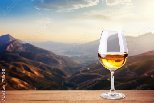A single glass of white wine placed on a wooden table with a scenic mountain valley backdrop during sunset.