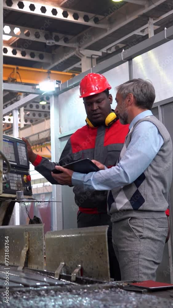 Production Manager Using Digital Tablet and Giving Instructions to African-American CNC Machine Operator. Supervisor Talking to CNC Machine Operator Next To Lathe Machine. Multiracial Coworkers. Production Manager Using Digital Tablet and Giving Instructions to African-American CNC Machine Operator. Supervisor Talking to CNC Machine Operator Next To Lathe Machine. Multiracial Coworkers.