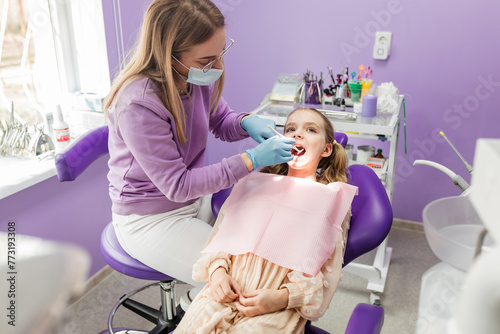 The dental surgeon makes the oral examination of mouth to female child in the dental office. The young girl sit in dental chair with open mouth during the doctor's examination