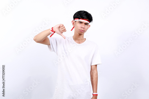 Unhappy asian young boy with red white ribbon celebrating Indonesia independence day while showing thumbs down photo