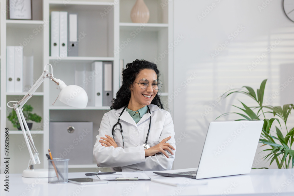 Portrait of a cheerful Hispanic female doctor wearing glasses, sitting in her clinic office with a laptop, emanating professionalism and care. Portrait of a cheerful Hispanic female doctor wearing glasses, sitting in her clinic office with a laptop, emanating professionalism and care.