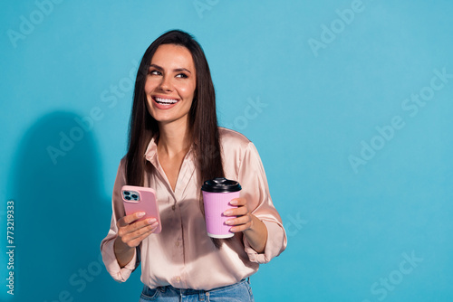 Photo of positive girl with straight hair wear silk blouse hold cup of latte smartphone look empty space isolated on blue color background