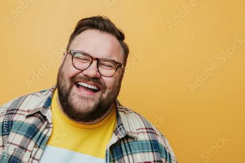 A bearded man with glasses is laughing heartily, dressed in a plaid shirt over a yellow tee, on a yellow background photo