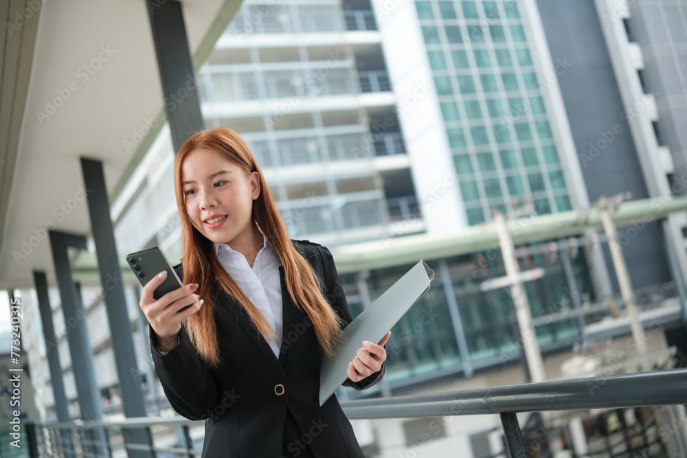 A young Asian businesswomen wearing a suit holding files standing in a big city on a busy downtown street. Young Asian businesswoman using smartphone texting to contact clients. A young Asian businesswomen wearing a suit holding files standing in a big city on a busy downtown street. Young Asian businesswoman using smartphone texting to contact clients.