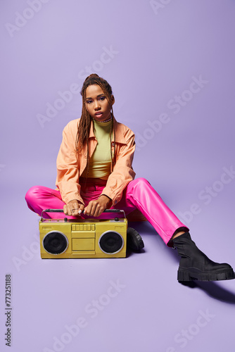 stylish african american woman with dreadlocks sitting next to retro boombox on purple backdrop