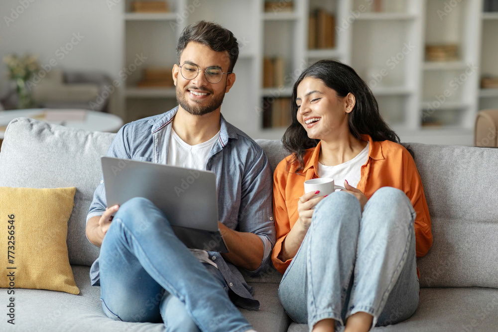 Indian spouses sitting on sofa in homey setting, shares a delightful moment browsing laptop together and smiling