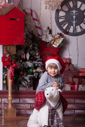 A portrait of a little girl in a red Santa hat is sitting on an artificial white bear (red, white and gray Christmas interior). Vertical image.
