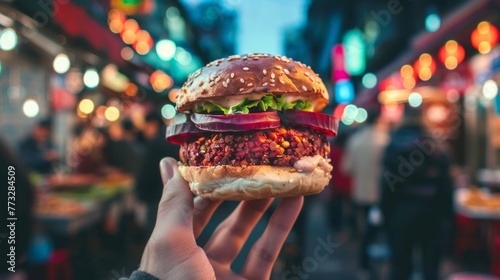 Hand holding a vegan burger with a colorful patty and fresh vegetables, with the lively ambiance of a street food market in the background. photo