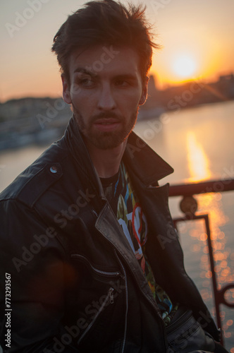 a brunette man in a leather jacket walks around the city at sunset photo
