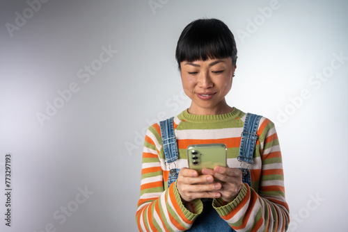 Smiling Asian woman in sweater posing isolated on grey wall background studio portrait. People lifestyle concept. Mock up copy space. Using mobile phone, typing sms message