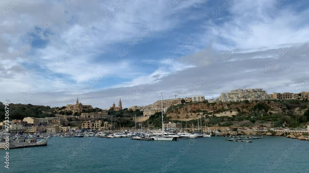Panoramic skyline view of Mgarr harbour and town. Yachts and boats at docks. POV view from a moving passengers ferry. Motion of sea waves. Mgarr, Gozo island, Malta Panoramic skyline view of Mgarr harbour and town. Yachts and boats at docks. POV view from a moving passengers ferry. Motion of sea waves. Mgarr, Gozo island, Malta
