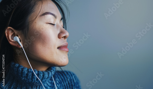 Young Japanese female model in a blue sweater with closed eyes enjoys listening to music on headphones, copy space for concept