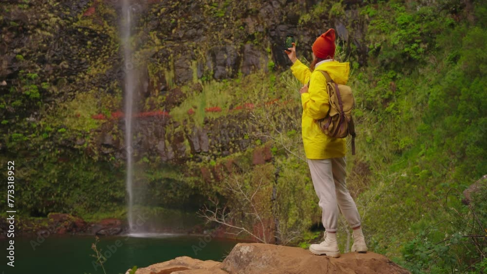A girl traveler in a yellow jacket and a backpack reached a beautiful, inaccessible waterfall and took pictures of it on her phone A girl traveler in a yellow jacket and a backpack reached a beautiful, inaccessible waterfall and took pictures of it on her phone