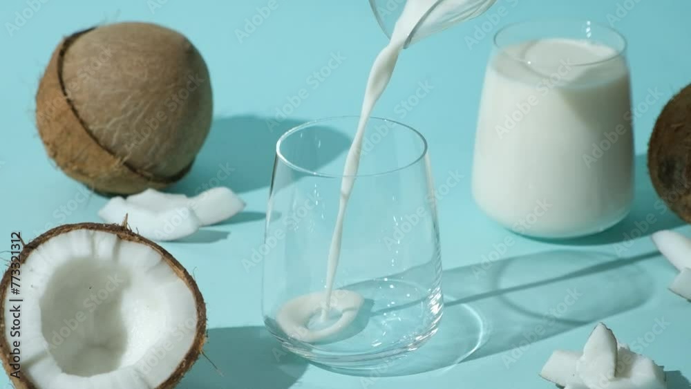 Coconut milk pouring into a glass among coconuts on a blue background, slow motion. Coconut milk pouring into a glass among coconuts on a blue background, slow motion.
