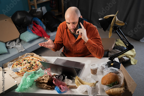 Slatternly bearded man sitting before laptop and holding smartphone among leftovers, food, bags and rubbish on background