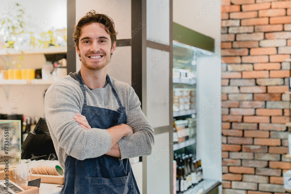 Fototapeta premium Smiling young white handsome barista standing with his arms crossed in coffee shop. Caucasian male bartender shop assistant vendor waiter looking at camera in cafeteria restaurant