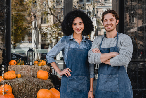 Portrait of young man and woman cafe owners waiters baristas bartenders service staff standing outdoor front of their cafe with decorating pumpkins preparing for Halloween.