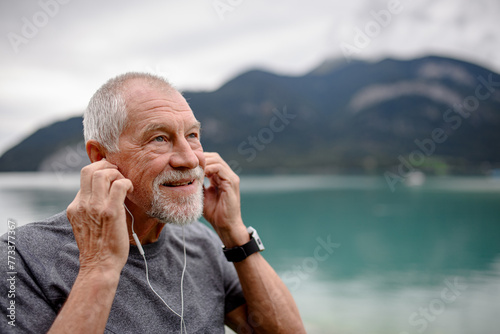 Senior listening music while running by lake in nature. Elderly man exercising to stay healthy, vital, enjoying physical activity and relaxation outdoors.